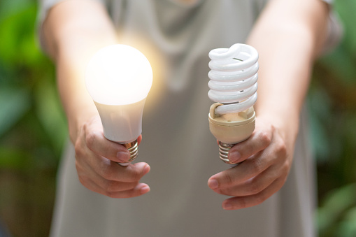 Power saving concept. Hands holding new Light Emitting Diode ( LED ) light bulb with light on and blur spiral compact-fluorescent (CFL) bulbs behind for copyspace.