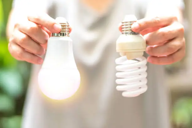Photo of Power saving concept. Hands holding new Light Emitting Diode ( LED ) light bulb with light on and blur spiral compact-fluorescent (CFL) bulbs behind for copyspace.