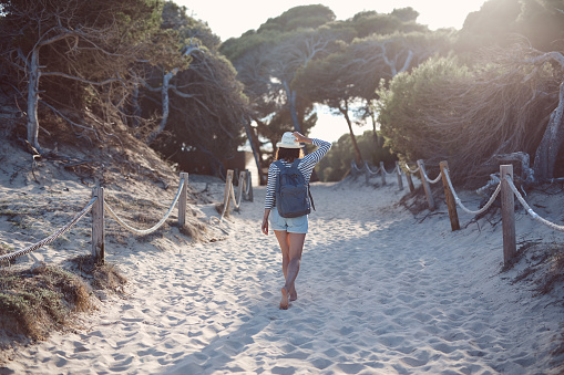 A photo of young woman walking on sandy footpath amidst plants in the summer. She is wearing backpack, holding her hat with a palm, backlit by setting sun.