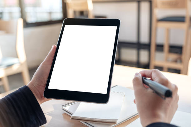 istock Mockup image of hands holding and using black tablet pc with blank white desktop screen while writing on notebooks in office 1058262510