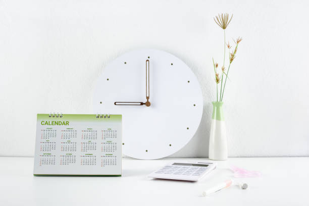 istock Clear, clean theme white office desk concept : Business working space and office supply on a table e.g flipped desktop calendar, analog clock, calculator, pen, scissor, two toned vase near white wall 1058276412