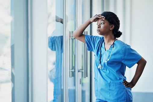 Shot of a young female nurse looking stressed out while standing at a window in a hospital