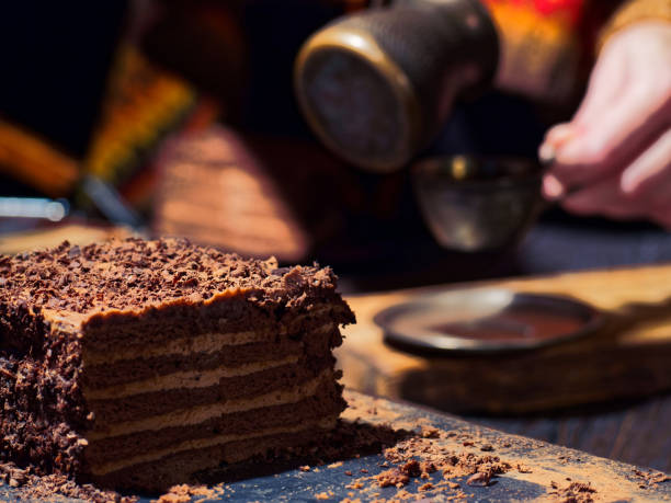 Homemade chocolate cake is on the foreground. A person wearing colorful woolen sweater is making a cup of coffee on the background.