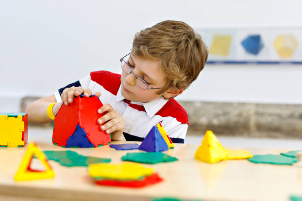 menino de garoto com óculos jogando com kit de elementos plásticos lolorful na escola ou creche pré-escolar. criança feliz construindo e criando figuras geométricas, aprendizagem matemática e geometria. - building activity block human hand house - fotografias e filmes do acervo