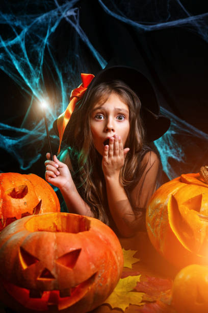 istock Halloween concept - little witch child enjoy playing with magic wand over spider web and with curved pumpkins background. 1058309186