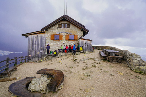Dolomites, Italy - September 14, 2018: Dolomites, Italy - September 14, 2018: View of tourists hiding from rain under the roof of The Rifugio Nuvolau mountain cabin at Averau-Nuvolau group in Dolomites, Italy. In background is dark, cloudy sky. The cottage is located on the same Nuvalou top, which is located southwest of Cortina d'Ampezzo. From the summit, in good weather, a beautiful view opens in the heart of the highest peaks of the Dolomites.