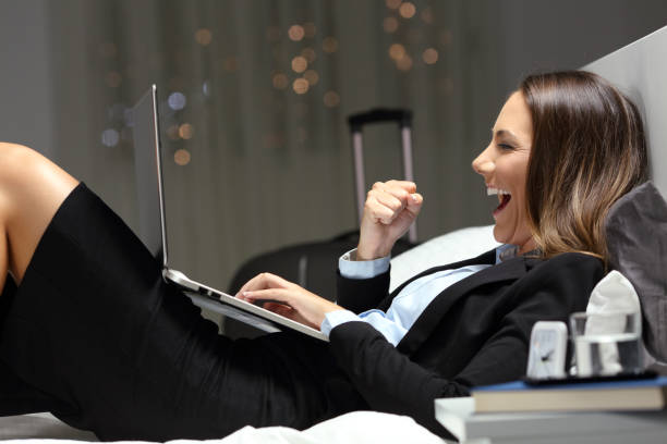 Excited bussinesswoman finding online content at hotel Excited bussinesswoman during a business travel finding online content in a laptop lying on a bed at hotel room travel refund stock pictures, royalty-free photos & images