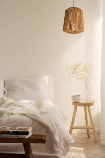 istock Bedside table with mug and flower next to bed with white bedding, pillow and blanket, real photo with copy space on the empty wall 1058365024
