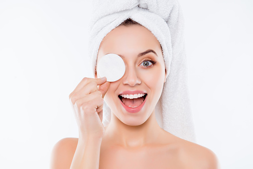 Portrait of foolish playful woman using sponge for application of lotion close one eye with cotton keeping open mouth with towel on head isolated on white background.