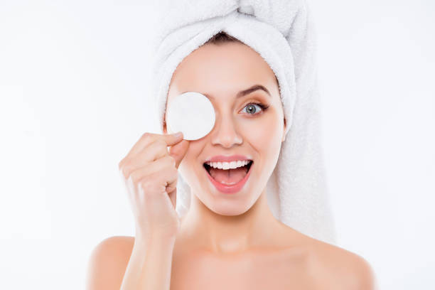 portrait of foolish playful woman using sponge for application of lotion close one eye with cotton keeping open mouth with towel on head isolated on white background. - facial cleanser imagens e fotografias de stock