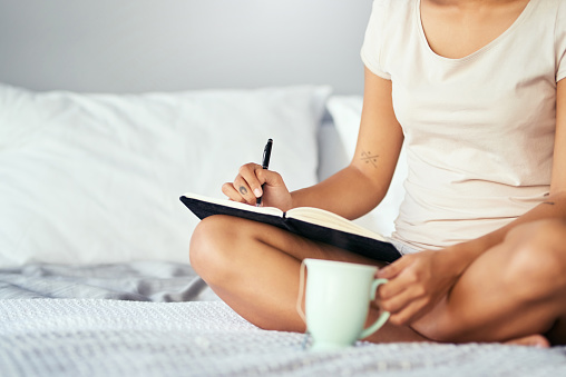 Cropped shot of a woman sitting on her bed and writing in a journal in the morning