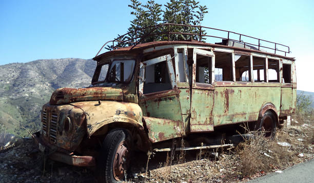 A rusty yellow mobile home rests in a vast, dry field, showcasing signs of age and neglect. Lush green forest mountains rise majestically in the background under a bright, sunny day.