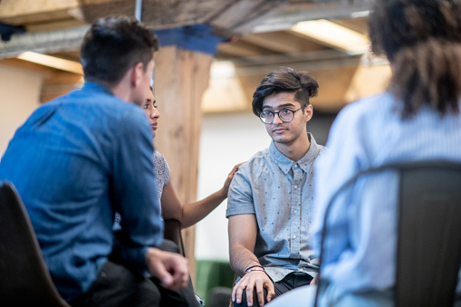 A man looks forlorn as he sits in a circle of young adults, who are looking at him. The woman next to him has her hand on his shoulder, offering him her sympathy.