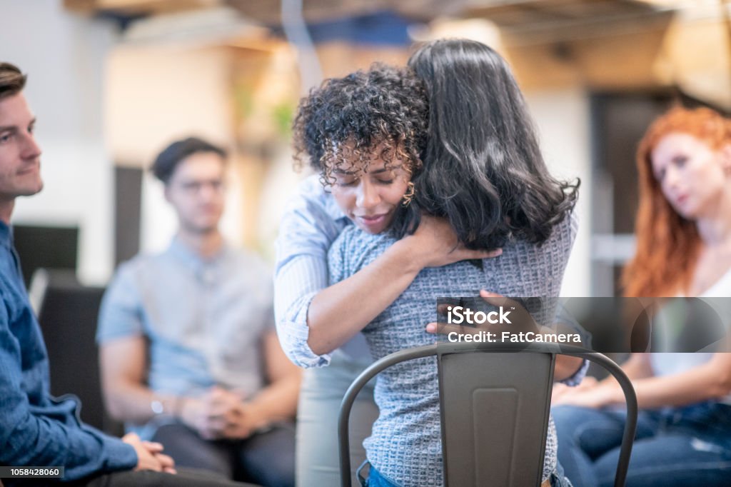 Two women hugging during a support group meeting A woman gives a genuine hug to another woman who is sitting in her chair, at a support group meeting where a diverse group of people are sitting in their chairs which are positioned in a circle. Group Therapy Stock Photo