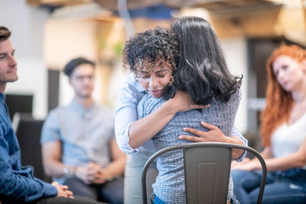 Two women hugging during a support group meeting A woman gives a genuine hug to another woman who is sitting in her chair, at a support group meeting where a diverse group of people are sitting in their chairs which are positioned in a circle. alcoholics anonymous photos stock pictures, royalty-free photos & images