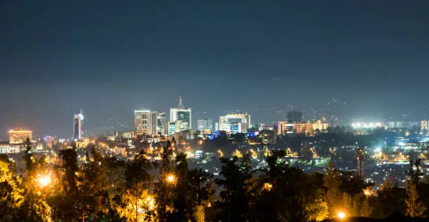 A panoramic view of Kigali city skyline lit up at night, under a deep blue evening sky with the glow of streetlights in the foreground