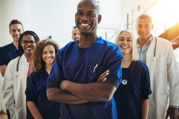 Smiling African doctor standing in a hospital with his staff Young African male doctor smiling while standing in a hospital corridor with a diverse group of staff in the background medical occupation stock pictures, royalty-free photos & images