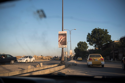 Several months after the eastern half of Mosul was liberated by the Iraqi government from ISIS, cars drive past a banner on a light pole that can be translated from the Arabic as \