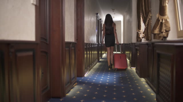 Back view of a brunette walking along the hotel corridor with the trolley suitcase. The camera moves bottom-up