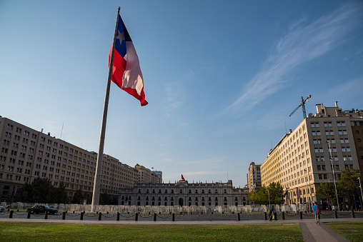 Santiago de Chile, Chile; February 04, 2018: La Moneda House of government of Chile with a giant flaming flag of Chile and a part of La Alameda street, a car passing by and two people walking