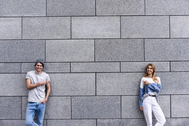 Photo of Handsome couple is pointing away and smiling, standing against gray wall.