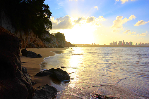 Cliffs, rocks, plants, beach, skyline of the city, blue sky with clouds, sunshine reflected by the flat sea
