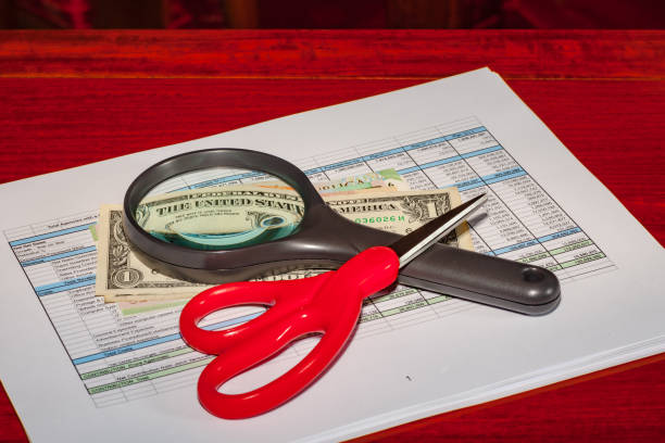 Budget tools - Instruction to the management team of the company to reduce business operational costs. The pair of scissors signifies the intention to cut operational costs (be wise); the magnifying glass, to look for savings and to find every dollar that can be saved (be innovative). The glass distorts the dollar. Spreadsheet prepared as a prop and has no real-life significance. Image shot in studio environment. Camera: Canon EOS 5D MII. Lens: Leica APO Summicron - R 90mm F2 ASPH. herramientas de presupuesto - imagen conceptual con un par de dólares, lupa y un par de tijeras con un presupuesto de la empresa, mandar el equipo figurativamente para reducir los costes operativos y ser innovadores con el presupuesto; imagen horizontal t