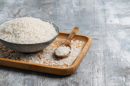 Raw white rice in ceramic bowl with wooden spoon over gray background. Wabi Sabi style. Copy space. Horizontal.