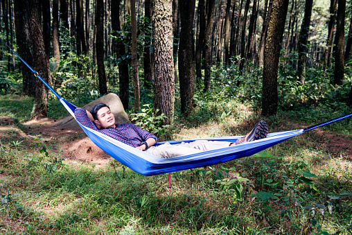 Young asian man relaxing on the hammock hanging among trees on the forest