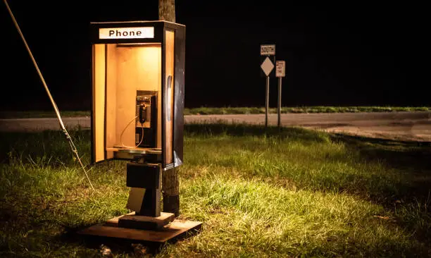 An old phone booth is lit up on the side of a rural highway at night.