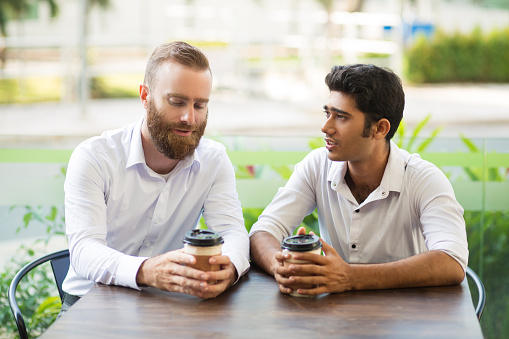 Two business men drinking coffee and chatting in outdoor cafe. People sitting at table with blurred plants in background. Coffee break concept. Front view.