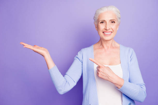 istock Grey haired old nice beautiful smiling woman holding product object, pointing with finger, copy blank empty space. Isolated over violet purple background 1058650956