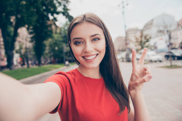 Self-portrait of straight-haired young beautiful smiling girl wearing casual red t-shirt outside, traveling abroad, showing v-sign Self-portrait of straight-haired young beautiful smiling girl wearing casual red t-shirt outside, traveling abroad, showing v-sign peace sign gesture photos stock pictures, royalty-free photos & images