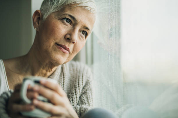 istock Thoughtful senior woman with tea cup looking through window during rainy day. 1058663452