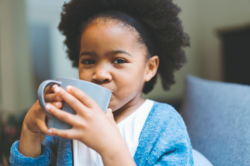 Portrait of cute girl drinking from mug at home. Close-up of female child is sitting in living room. She is with afro hairstyle.