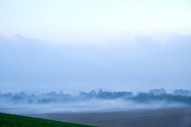 British countryside looking across a sweeping field to an old english village covered in white morning mist, the trees and the buildings are above the mist and the church tower can be seen silhoutted by the mist above the mist is grey sky, Firle village, Sussex, England