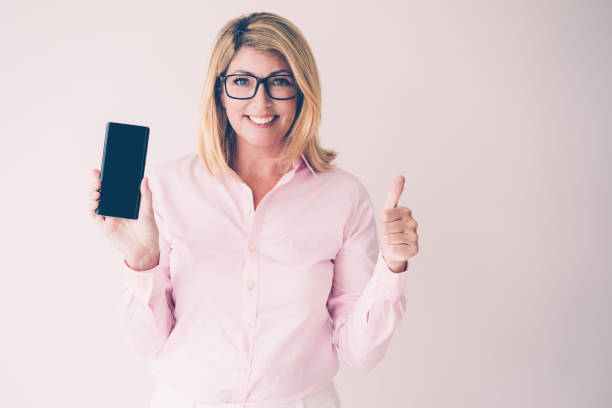 Closeup portrait of smiling middle-aged attractive fair-haired woman showing thumb up and smartphone screen. Technology concept. Isolated front view on grey background.