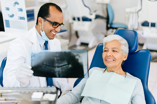 A cheerful male dentist sitting next to a senior woman patient, holding and showing her an x-ray of her teeth.
