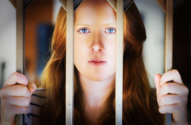 A young woman looks seriously at camera from behind bars which may be prison bars or those of a security gate.
