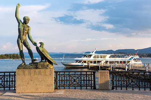 Zurich, Switzerland - August 19, 2018: MS Limmat approaching a pier on Lake Zurich at sunset, sculpture of Ganymede on the embankment of the lake, people on the pier, summits of the Alps in the background. MS Limmat is a ship of the Lake Zurich Navigation Company (German: Zurichsee-Schifffahrtsgesellschaft or ZSG), which is a public Swiss company operating passenger ships and boats on Lake Zurich.
