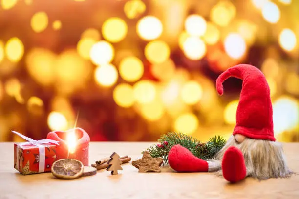 Red sitting christmas elf with bokeh lights in background surrounded by christmas decorations as gift box and ignited advent candle