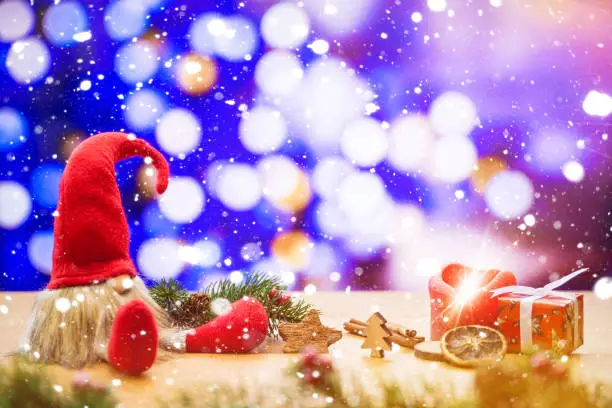 Red sitting christmas elf in falling snow with bokeh lights in background surrounded by christmas decorations as gift box and ignited advent candle