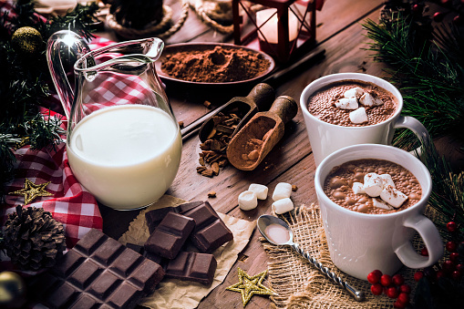 Homemade hot chocolate ingredients with mugs, marshmallows, cocoa, milk, sugar, cinnamon and chocolate bars on rustic wooden table with candlelight. Christmas themes.