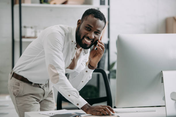 istock handsome african american businessman talking by smartphone and using computer in office, looking at camera 1058759792