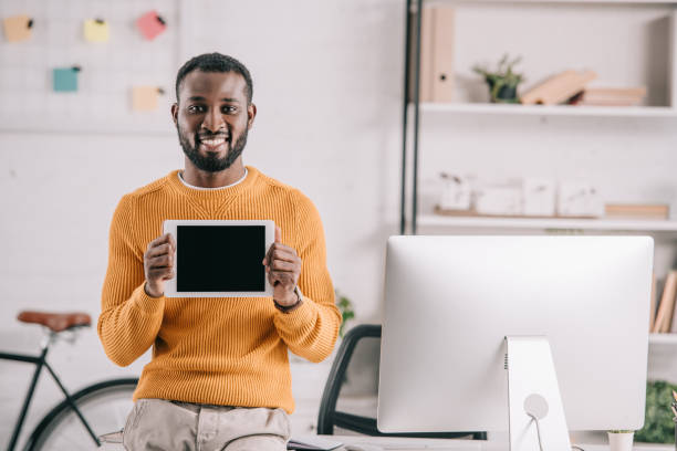 istock smiling handsome african american designer in orange sweater showing tablet with blank screen in office 1058770326