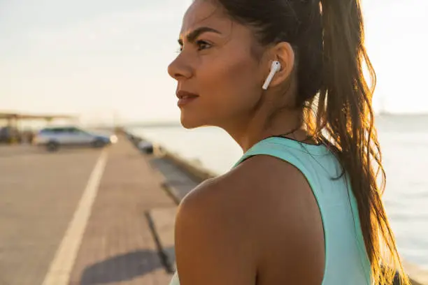 Tired fitness woman sweating taking a break listening to music on phone after difficult training. Young woman listening to music with earphones on smart phone app for fitness motivation