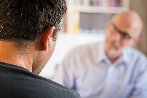 Mature male psychotherapist or counsellor interacts with a male client during a one-to-one therapy session.