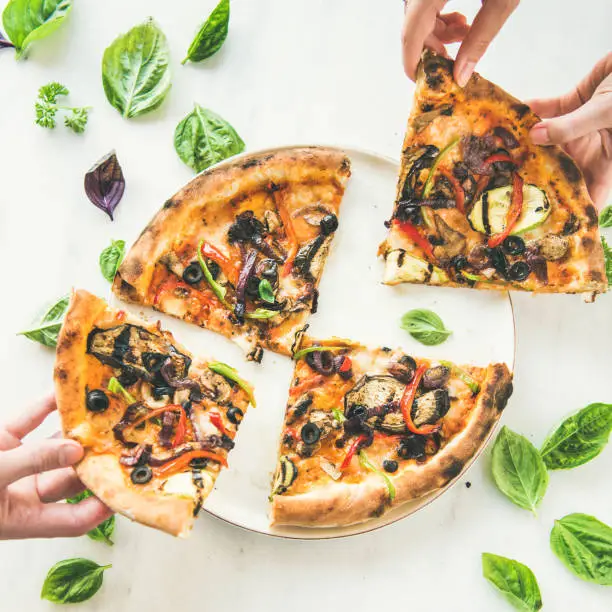 Summer dinner or lunch. Flat-lay of people's hands taking freshly baked Italian vegetarian pizza with vegetables and fresh basil over white marble table, top view, square crop