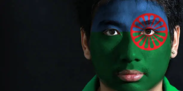 Portrait of a man with the flag of the Romani people painted on his face on black background. blue and green background, representing the heavens and earth and red chakra or cartwheel in the center.