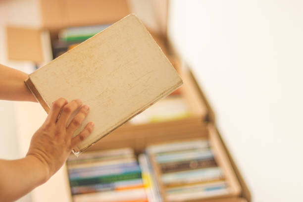 Female hand holding an old book. Defocused cardboard boxes full of books on background. Concepts of second hand bookstore and moving home.